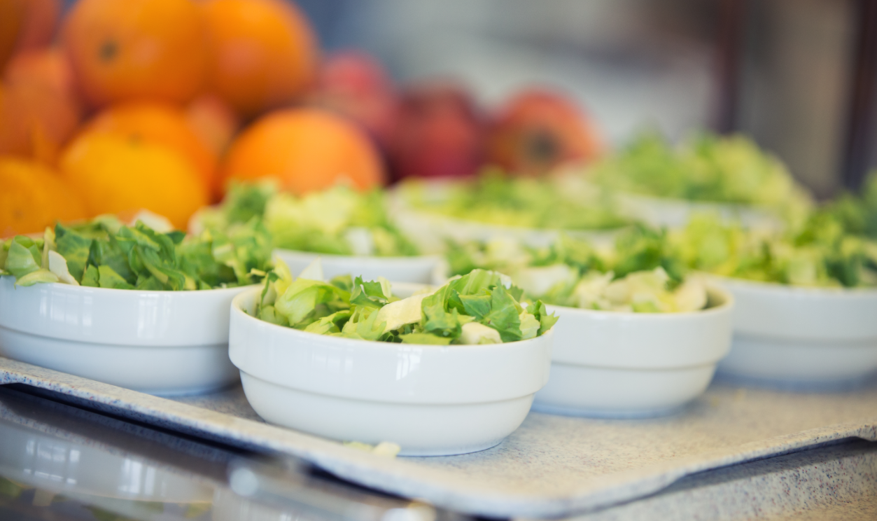 Ein Tablett mit kleinen Schalen voll grünem Salat. Im Hintergrund verschwommen: Orangen und Äpfel