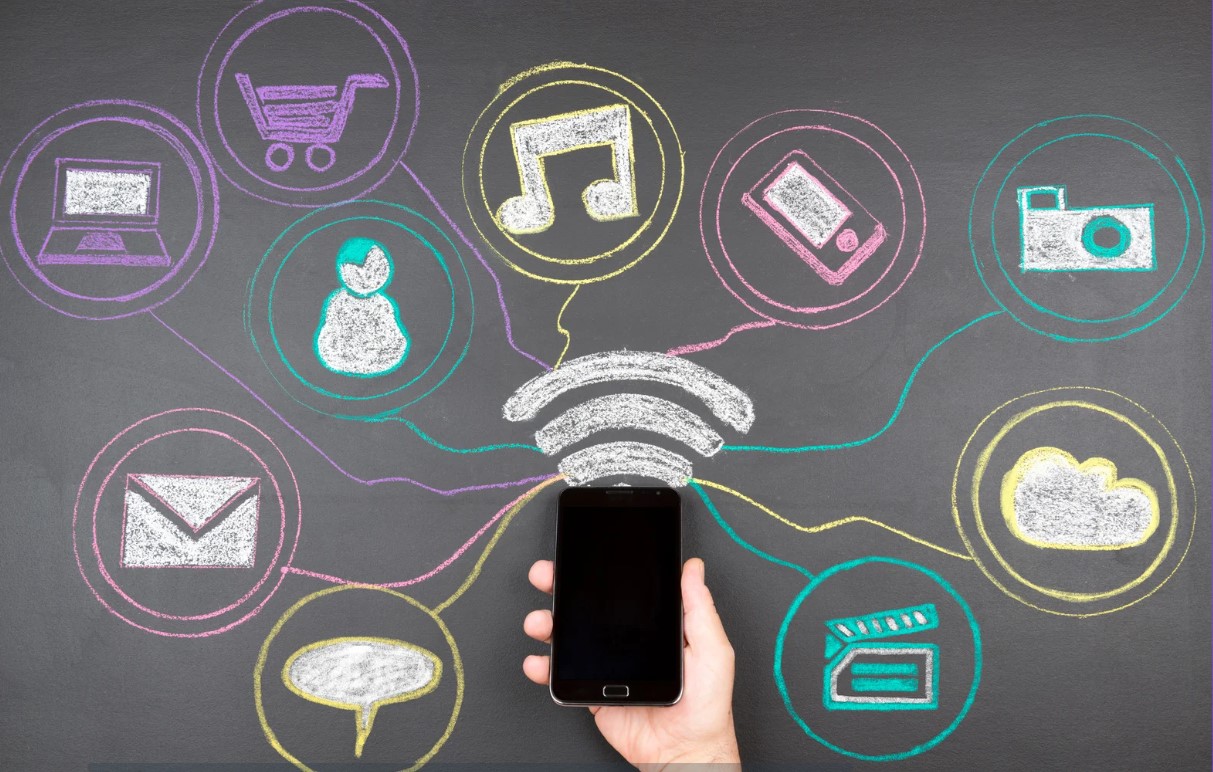 Eine Hand hält ein Smartphone. Davon ausgehend ist in Kreideoptik eine Sammlung verschiedener Symbole dargestellt. Zu sehen sind die Symbole Sprechblase, Briefumschlag, Mensch, Computer, Einkaufswagen, Musiknote, Smartphone, Kamera, Wolke und Film-Klappe.