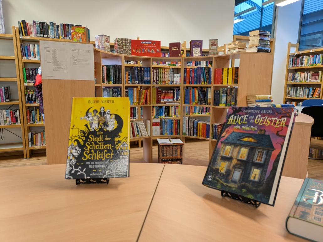 Foto der Schulbibliothek. Im Hintergrund: Bücherregale, im Vordergrund: Die Bücher Die Stadt der Schatten-Schläfer und Alice und die Geister von nebenan.