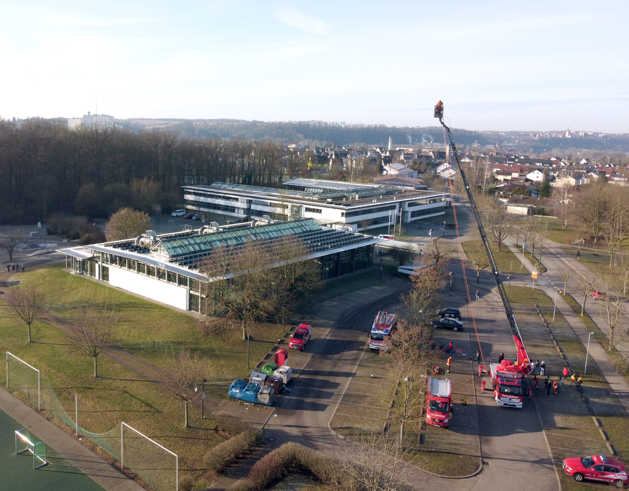Luftaufnahme des Parkplatzes vor der Schule. Im Vordergrund: Einsatzfahrzeuge der Feuerwehr. Die Drehleiter ist ganz ausgefahren. Im Hintergrund: Sporthalle und Schule