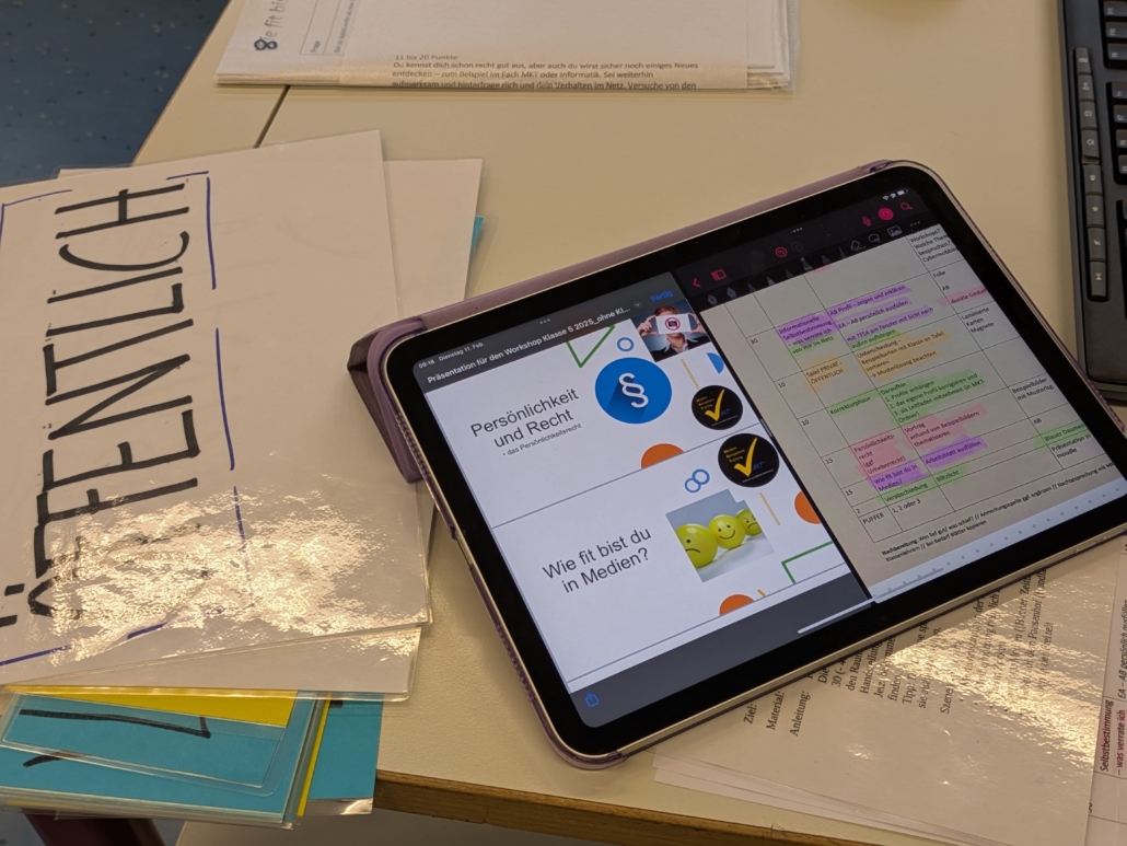 Foto eines Schultischs, auf dem laminierte Blätter liegen. Auf den blättern liegt ein Tablet-Computer, auf dem eine farblich markierte Tabelle erkennbar sind und zwei Rechtecke, in denen "Persönlichkeit und Recht" sowie "Wie fit bist du mit Medien?" zu lesen sind