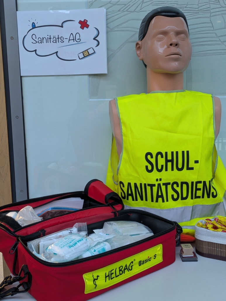 Oben links: Schriftzug Sanitäts-AG, rechts eine Reanimationspuppe mit einer gelben Schutzweste mit dem Schriftzug "Schulsanitätsdienst". Vorne links im Bild liegt eine geöffnete Sanitätstasche mit Verbandsmaterial - Aufschrift Helbag Basic S.