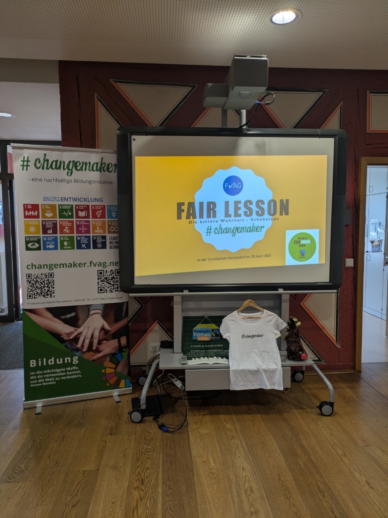 Zu sehen ist ein Aufsteller mit den Aufdrucken #changemaker und Bildung sowie zwei QR-Codes und den Nachhaltigkeitszielen. Daneben steht eine Projektionsfläche, auf der das Schullogo FvAG, der Schriftzug "Fair Lesson" und #changemaker erkennbar sind.