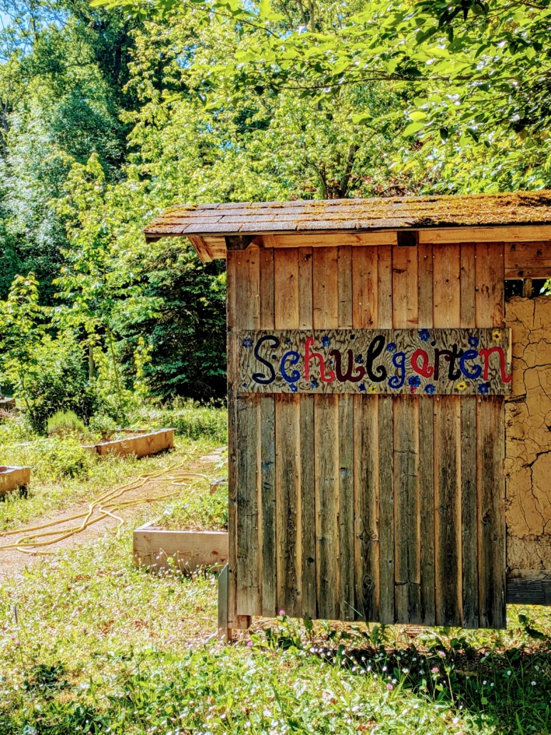 Foto des Schulgartens. Im Vordergrund: Insektenhoten mit der Aufschrift "Schulgarten", im Hintergrund Beete und ein Wasserschlauch.
