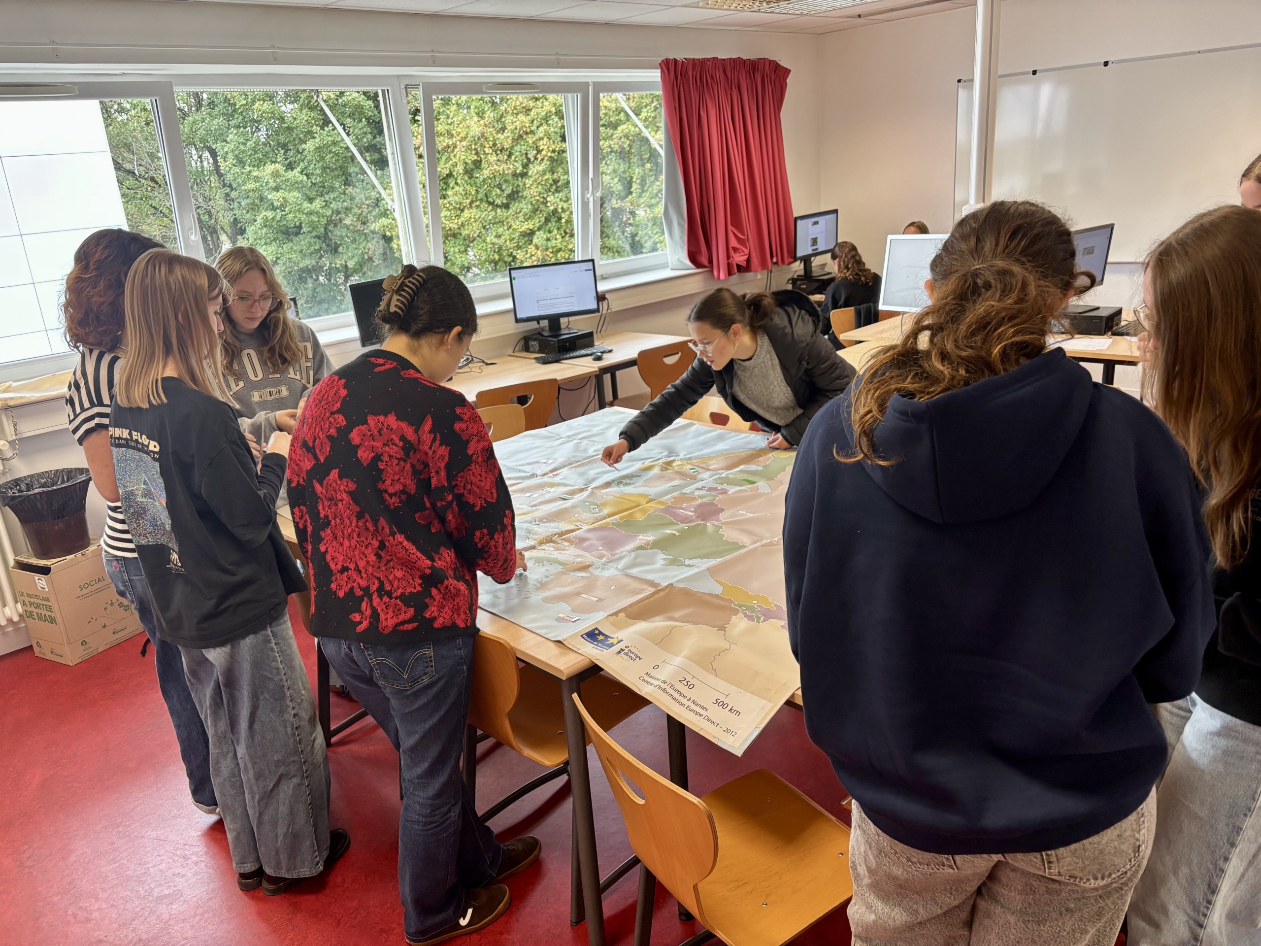 Foto von Schülergruppe bei einer Gruppenarbeit. In der Mitte steht ein großer Tisch mit einer Karte. Im Hintergrund ist eine Fensterfront erkennbar. Die Schüler stehen um den Tisch herum und schauen auf die Karte.