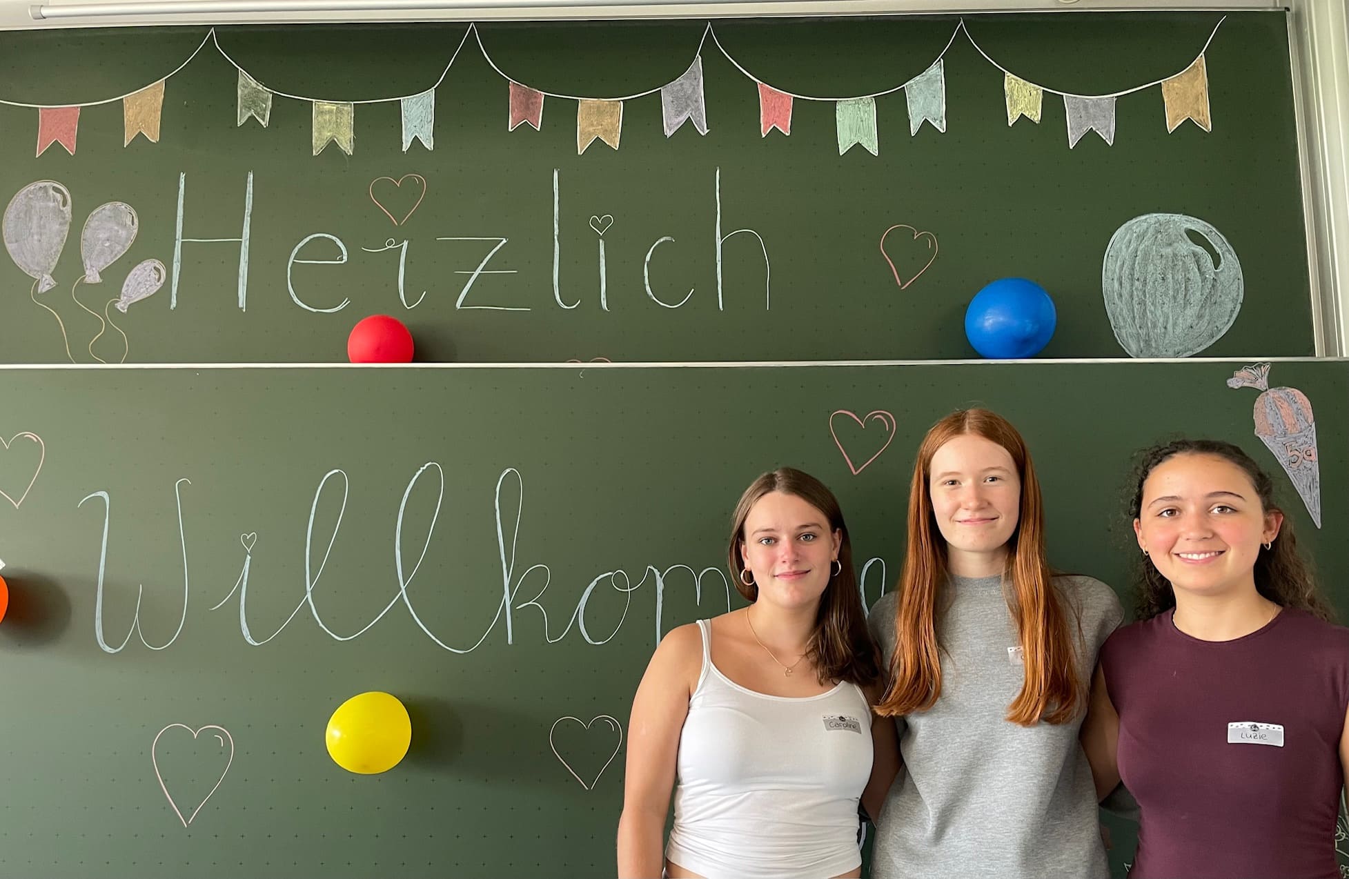 Drei Patinnen stehen vor einer bemalten Tafel. An dieser steht "Herzlich Willkommen"