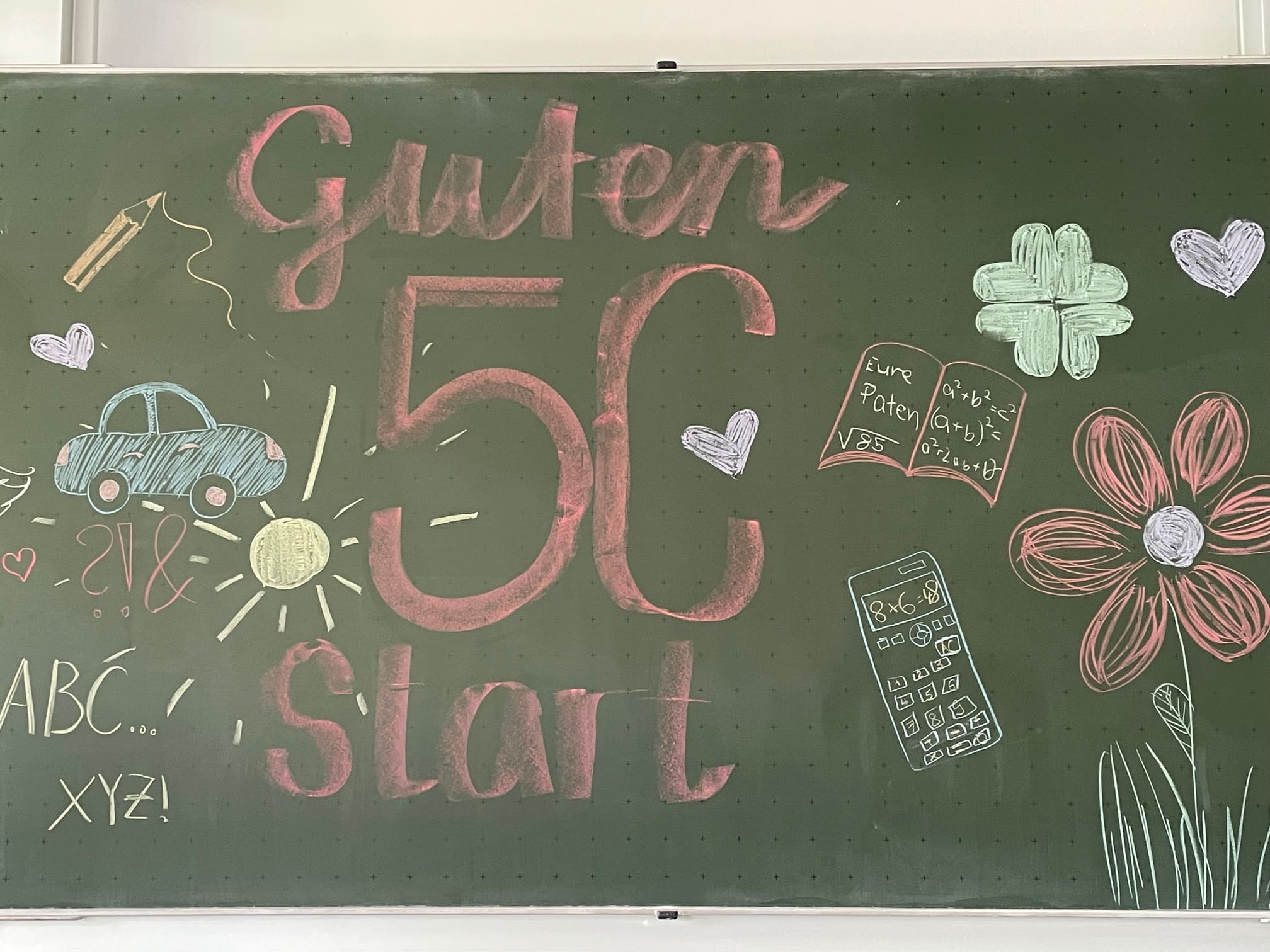 An einer bunt bemalten Tafel mit Blumen, Schulbüchern, einem Auto und einer Sonne steht "Guten Start 5c".