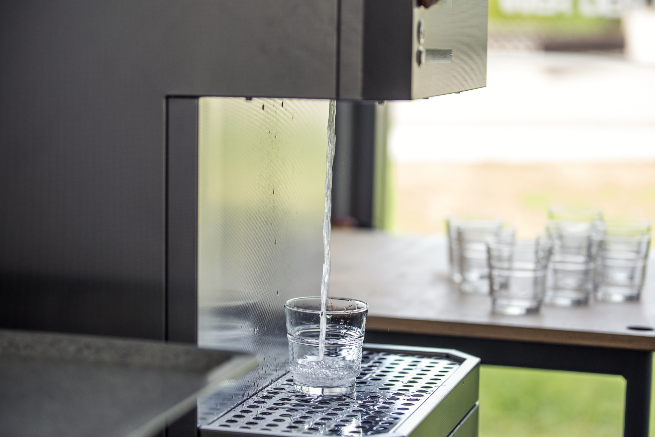 Foto eines Wasserspenders, aus dem Wasser in ein Glas fließt. Im Hintergrund stehen weitere Gläser.