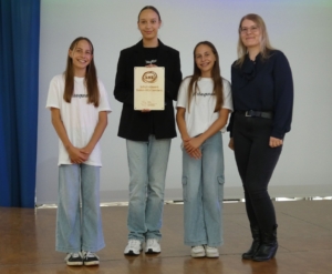 Nachhaltigkeitstag 25 (80) Drei Schülerinnen und Katharina Hauser stehen auf der Bühne in der Aula vor einer weißen Leinwand. Eine Schülerin hält das BNE-Siegel in der Hand.