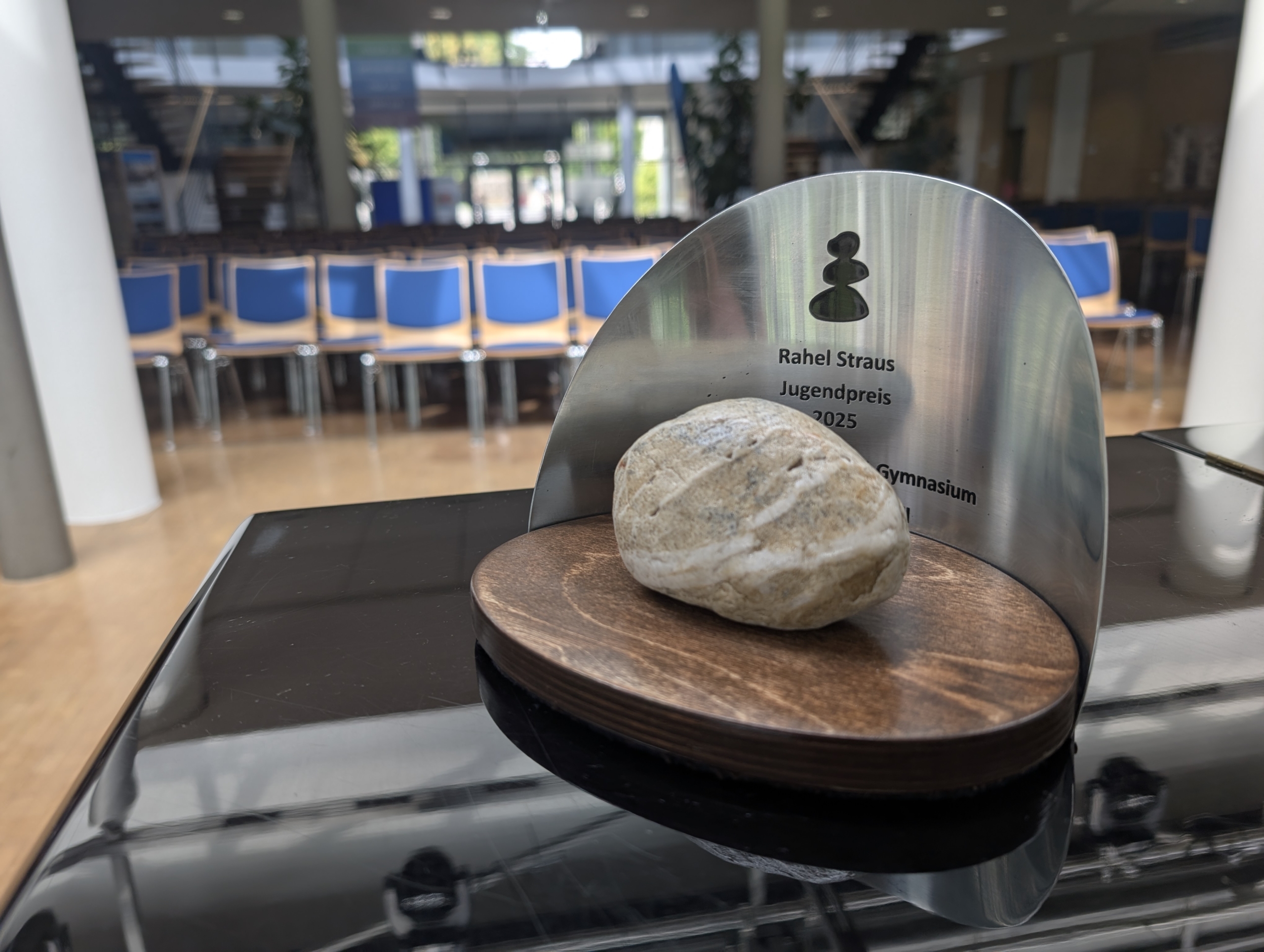 Der Rahel Straus Jugendpreis steht auf einem Flügel in der Aula der Schule. Im Hintergrund sind blaue Stühle erkennbar. Der Preis selbst besteht aus einem Stein auf einer Holzplatte. Dahinter befindet sich ein Metallschild mit der Aufschrift Rahel Straus Jugendpreis 2025.