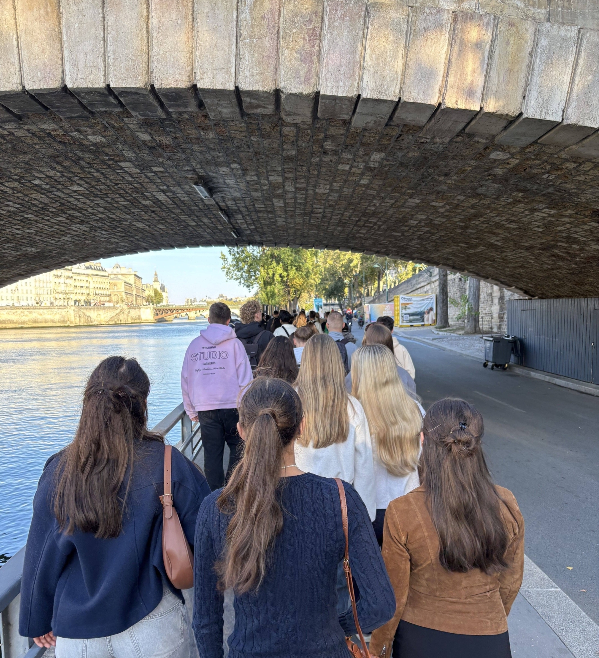 Eine Gruppe Schülerinnen und Schüler spaziert an der Seine in Paris entlang. Man sieht die Hinterköpfe der Personen. Oben ist eine Brücke erkennbar, links die Seine und rechts eine Straße.