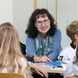 Foto von Fr. Smolka, die sich auf einen Schultisch lehnt, an dem vier Schüler sitzen. Auf dem Tisch liegen Unterrichtsmaterialien. Im Hintergrund ist Wanddekoration des Klassenzimmers erkennbar.