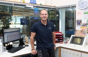 Foto von Hr. Stahl. Im Hintergrund ist sein Büro mit Monitoren, Büromaterial, eine Uhr, Fotos und eine große Glasscheibe erkennbar.