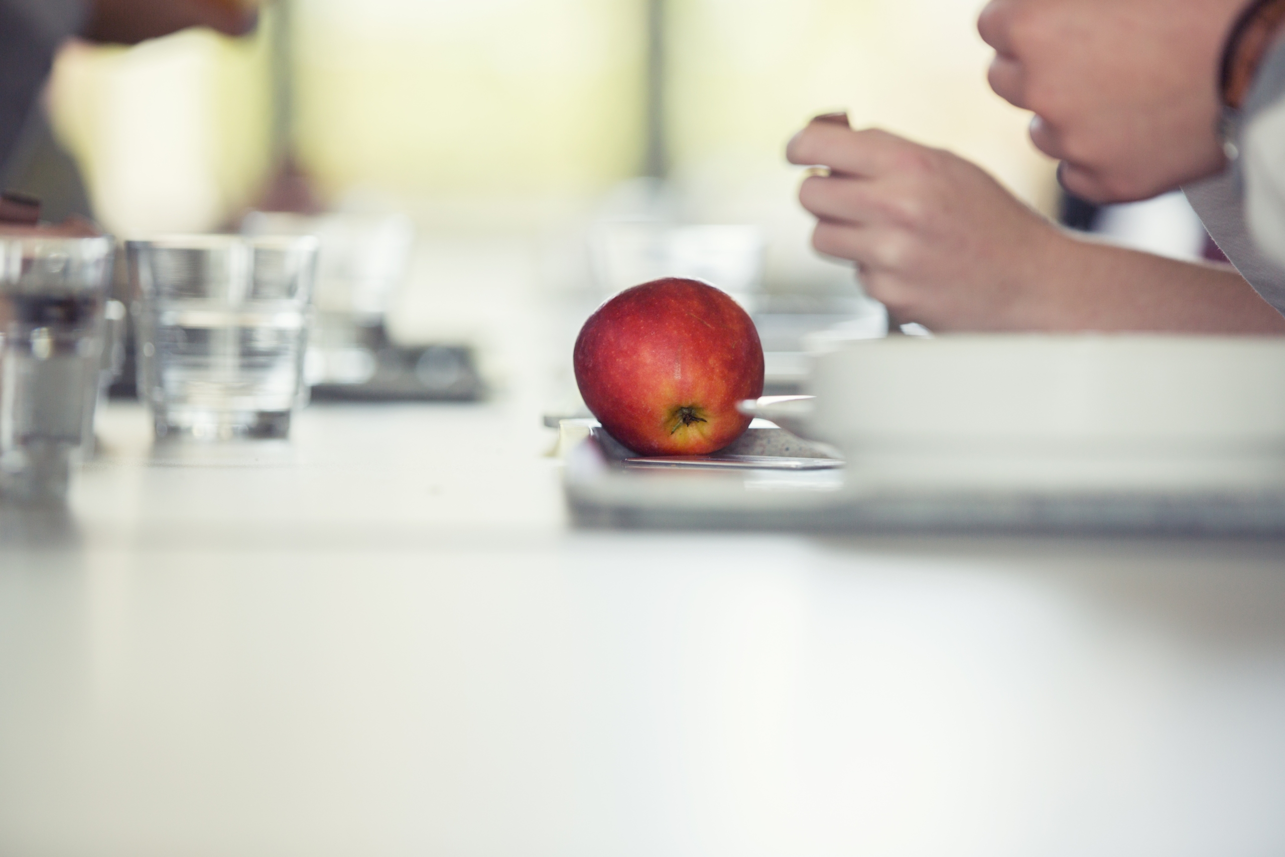 Unscharfe Aufnahme eines Tischs in der Mensa. Zu sehen sind Tabletts mit Gläsern und Tellern. Der Fokus liegt auf einem roten Apfel.