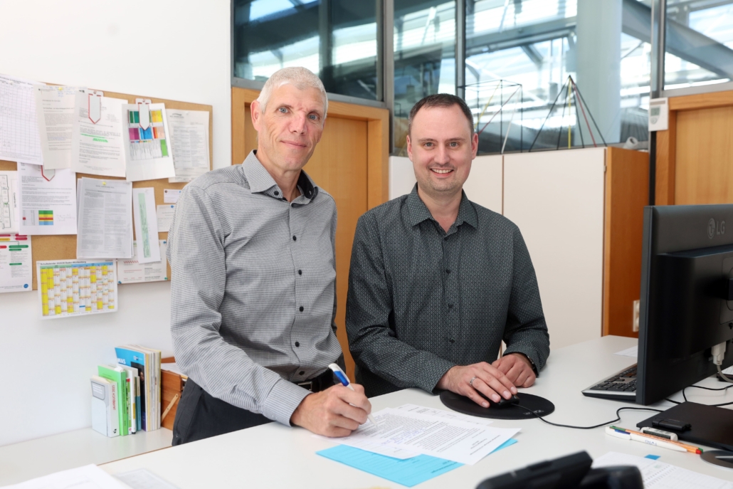 Foto der Oberstufenberater, die an einem Stehschreibtisch mit Computer stehen. Im Hintergrund ist eine Pinnwand mit vielen Papieren zu sehen. Außerdem im Hintergrund: Oberlichter, zwei Türen und ein Schrank.
