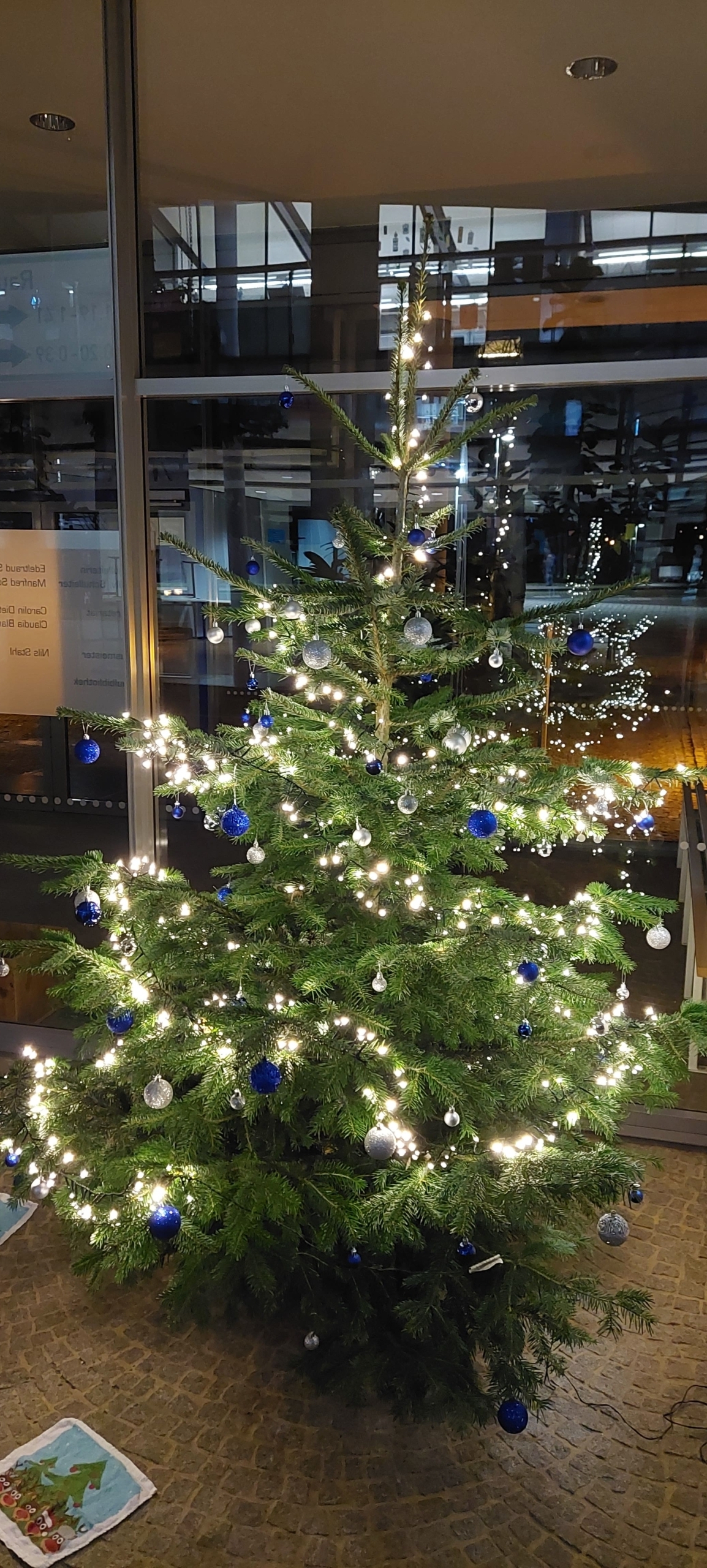 Abgebildet ist ein Tannenbaum im Foyer, der beleuchtet ist und mit blau-silbernen Kugeln dekoriert wurde.