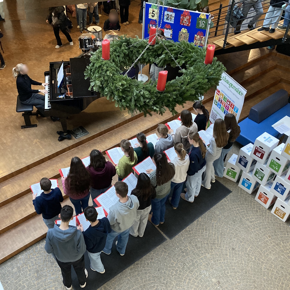 Schüler singen im Foyer, jemand spielt auf dem Flügel und der Adventskranz ist zu sehen.