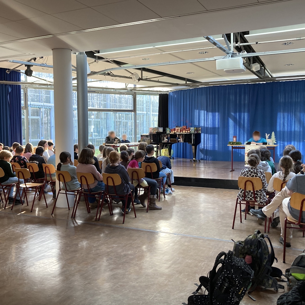 Schüler sitzen in der Aula und hören einem Schüler beim Vorlesen zu.