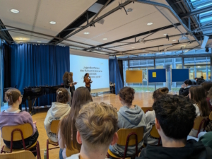 Schüler sitzen in der Aula, jemand präsentiert etwas.