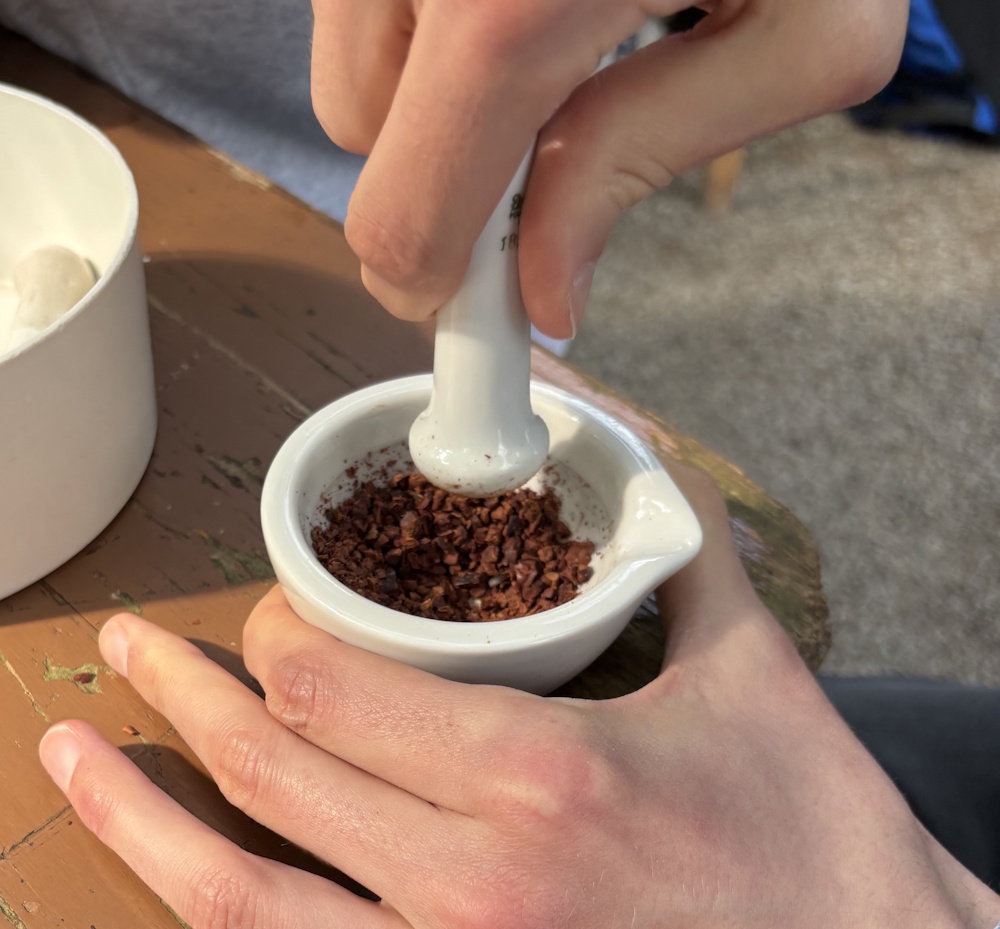 Zubereitung von Schokolade. Eine Hand zerstößt Kakaobohnen in einem Mörser.