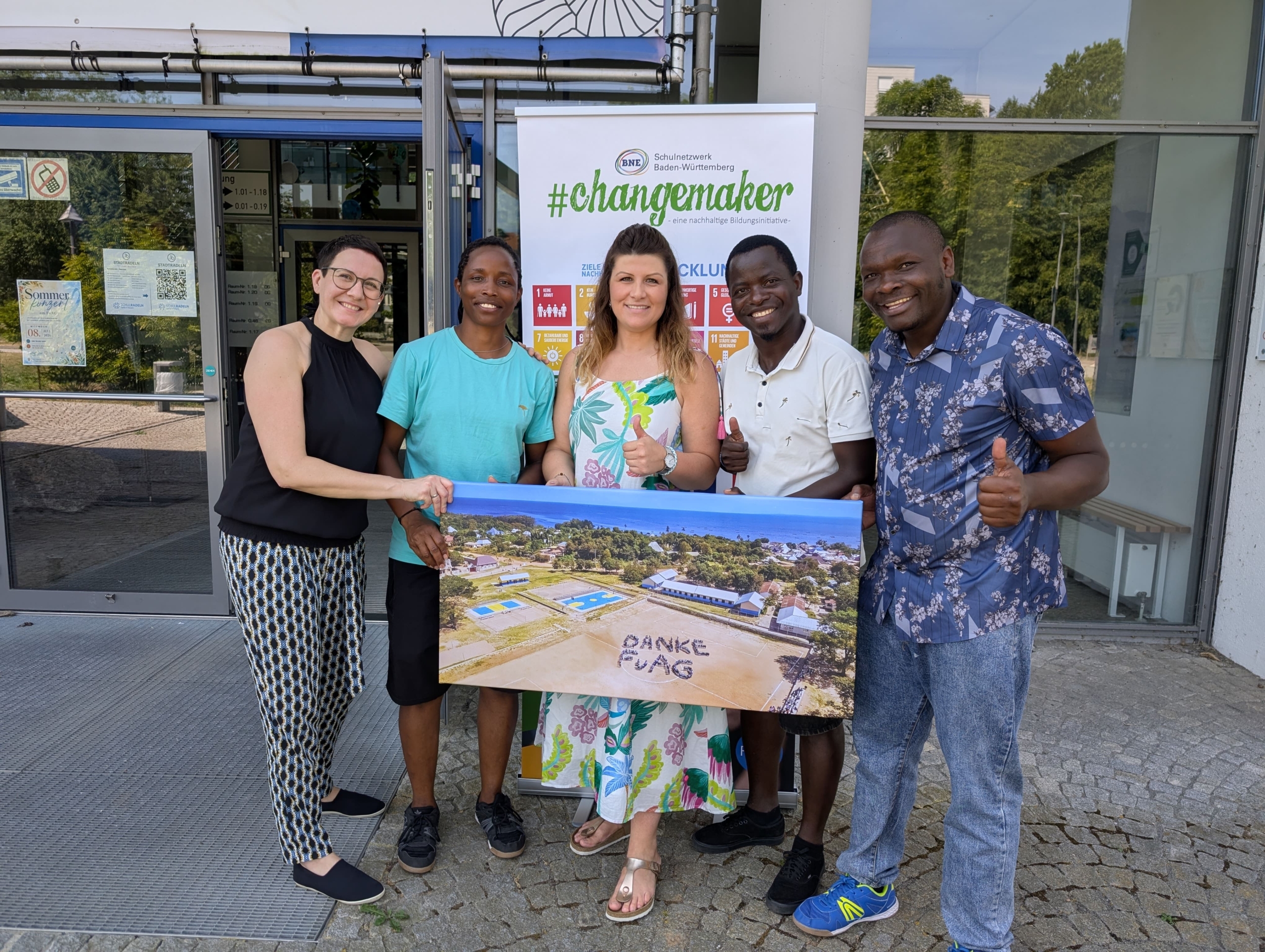 Zwei Lehrerinnen aus Deutschland und drei Lehrer aus Tansania stehen vor der Schule. Sie halten ein Bild vor sich, auf dem ein Sportplatz zu sehen ist. Darauf stehen Schüler, die den Schriftzug DANKE FvAG gebildet haben.